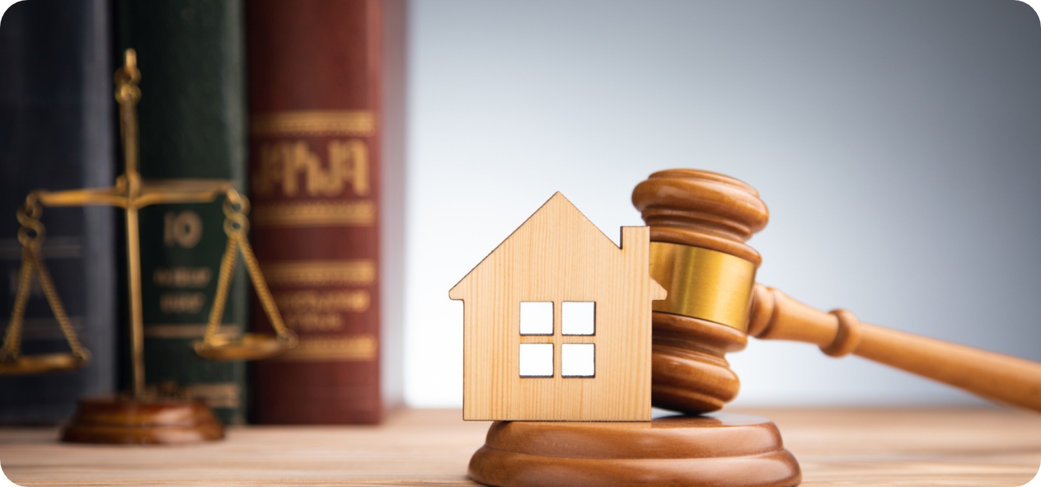 Richterhammer mit kleinem Holzhaus im Vordergrund und Büchern mit Gesetzestexten und Waage im Hintergrund.