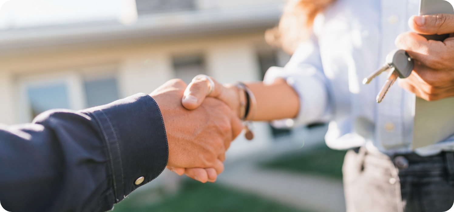 Zwei Personen, die einander die Hände schütteln, während eine Dokumente und einen Schlüssel hält.