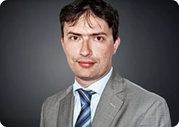 Porträt eines Mannes in einem grauen Anzug mit blauer Krawatte vor einem dunklen Hintergrund.