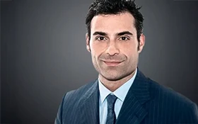 Porträt eines Mannes mit dunklem Haar, der einen blauen Anzug und eine hellblaue Krawatte trägt.