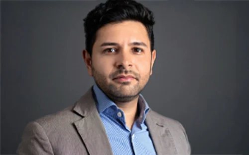 Porträt eines Mannes mit dunklen Haaren, der einen grauen Anzug und ein blaues Hemd trägt.