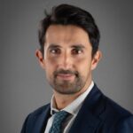 Mann mit dunklem Haar und Bart, trägt einen Anzug und eine Krawatte, posiert vor einem neutralen Hintergrund.