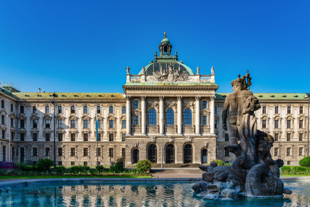 Vor der Münchner Residenz erhebt sich eine barocke Schlossfassade mit Brunnen und Skulptur unter klarem blauem Himmel.