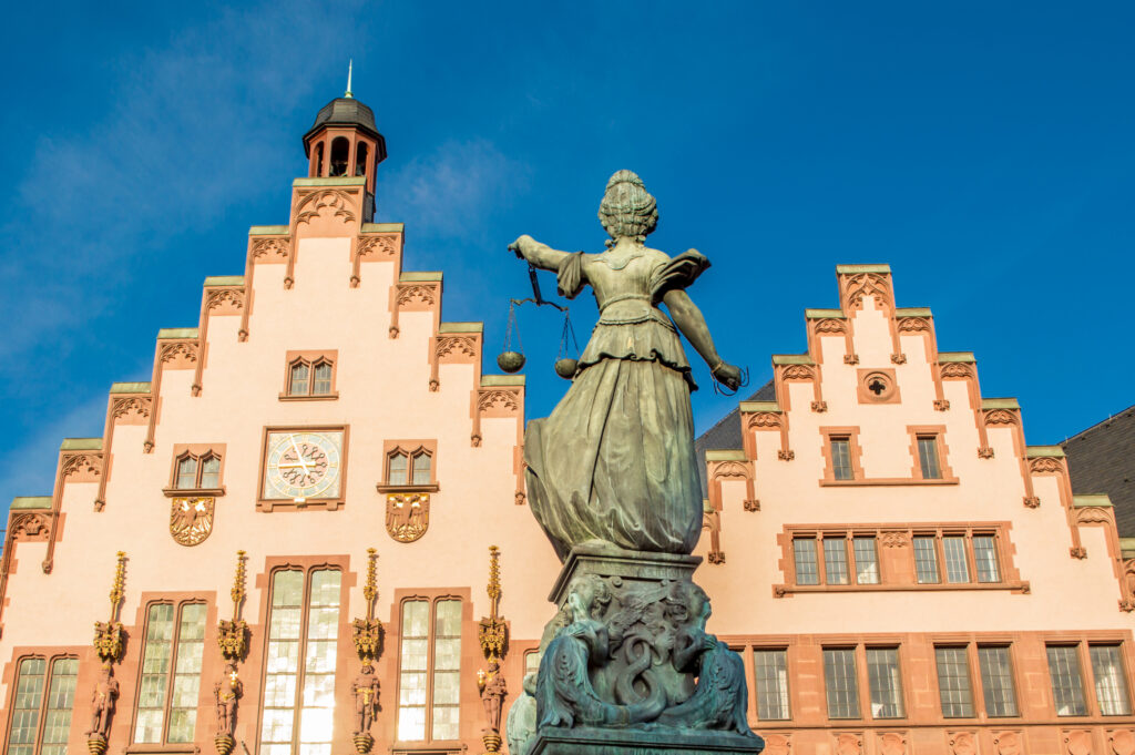 Vor dem Frankfurter Römer steht die Justitia-Statue vor der historischen Fassade mit Giebeln und Uhr unter blauem Himmel.